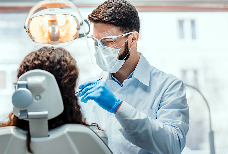 A dental professional in a white lab coat and blue gloves is performing a procedure on a patient s teeth, with the patient seated in a dental chair under bright overhead lights.