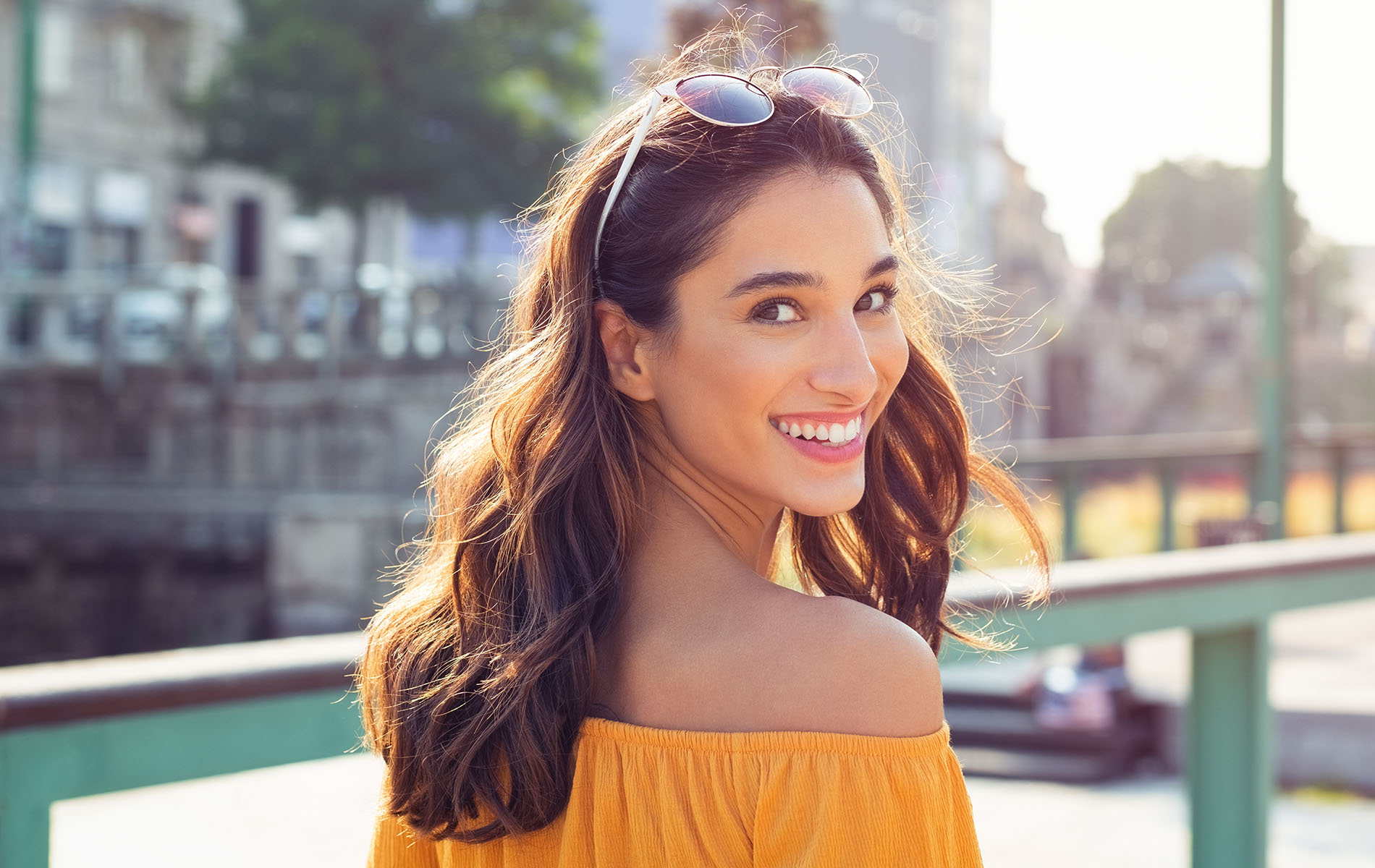 A woman with a radiant smile, wearing a yellow top and sunglasses, stands against a backdrop of an urban setting.