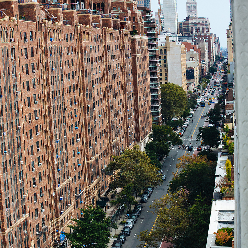 The image shows a city street with high-rise apartment buildings on both sides, a paved roadway with parked cars and traffic, trees lining the sidewalks, and a clear sky above.