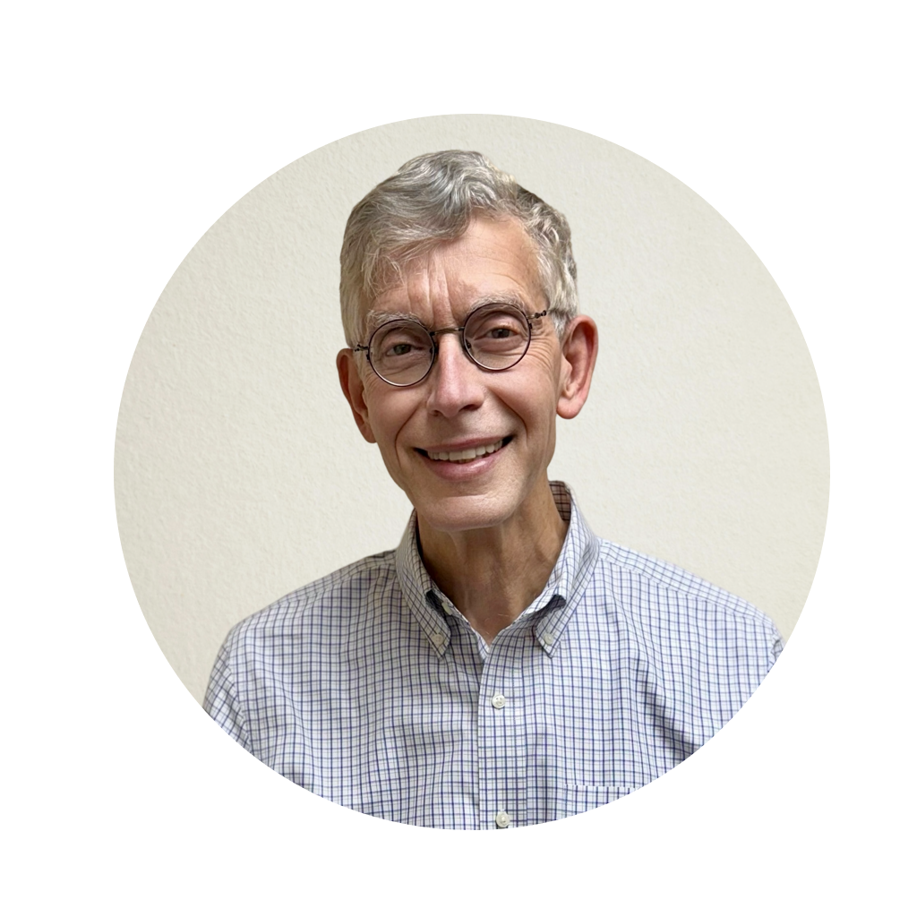 The image is a portrait of an older man with glasses, wearing a blue shirt and a necklace, posing for the photo. He has white hair and a beard, and he s smiling at the camera.