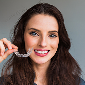 The image shows a woman holding up a transparent orthodontic appliance, possibly an aligner or clear retainer, with a smile.