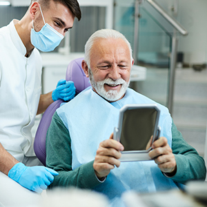 In the image, there is an older man sitting in a dental chair with a smile on his face while holding up a mirror to show his teeth. A dentist, wearing protective gear and a stethoscope, stands behind him, smiling at the camera. The setting appears to be a modern dental office, indicated by the presence of dental equipment and a professional atmosphere.