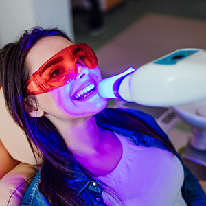 A woman sitting in a dental chair with purple lighting, wearing red goggles and having her teeth cleaned by a device.